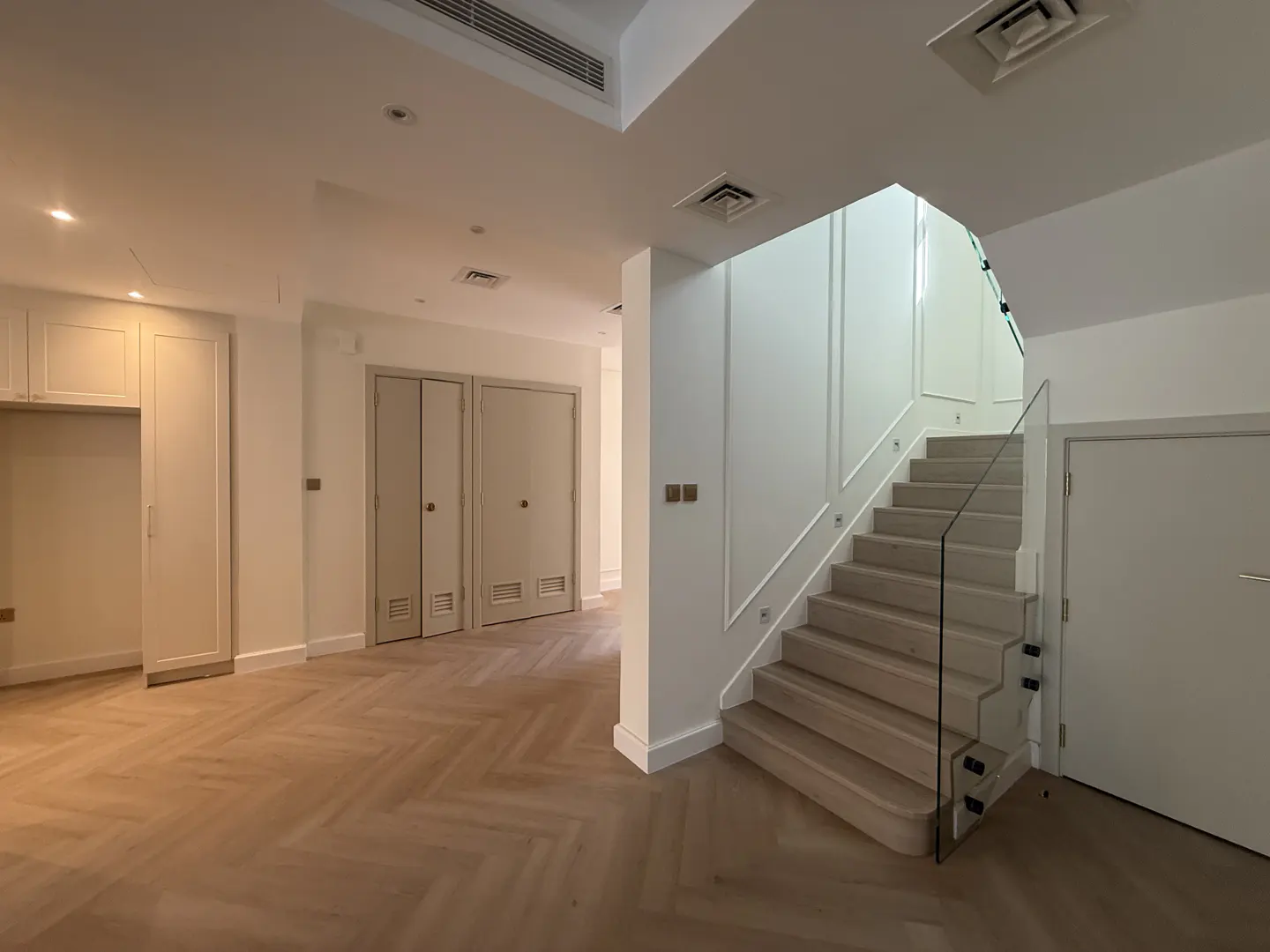 Bright, modern home interior with herringbone wood floors, white walls, built-in cabinets, and a staircase with glass railing.