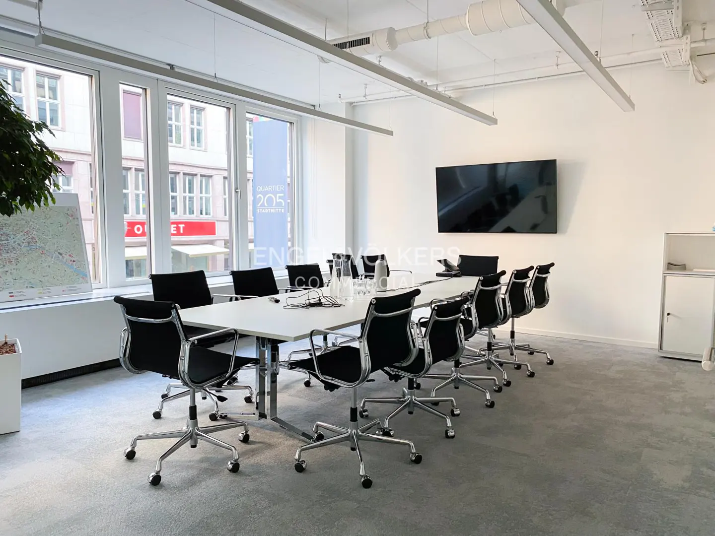 Bright, modern conference room with a long white table, black chairs, and a large screen TV on the wall. Large windows show a city view.