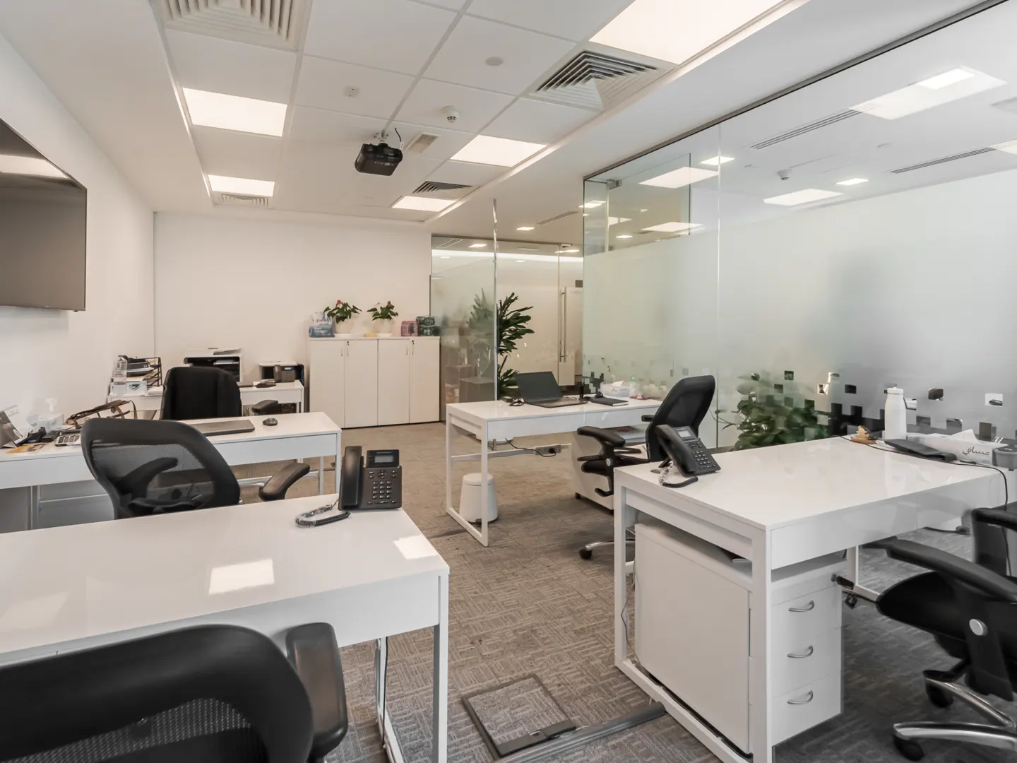 Modern office space with white desks, black ergonomic chairs, grey carpet, and glass partitions.