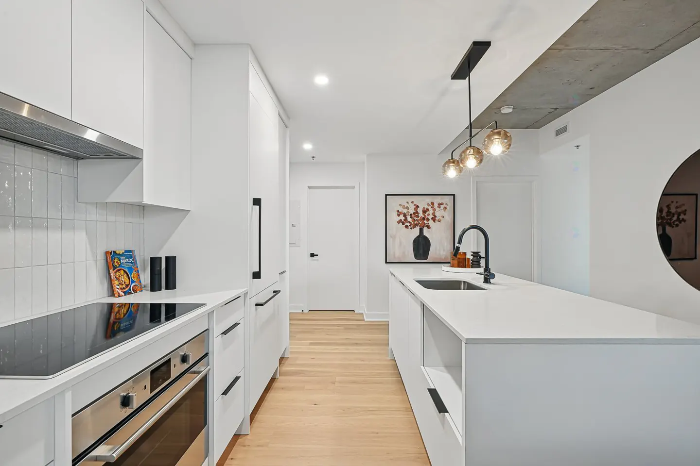 Modern white kitchen with light wood floors, a central island, black fixtures, and a concrete ceiling accent.