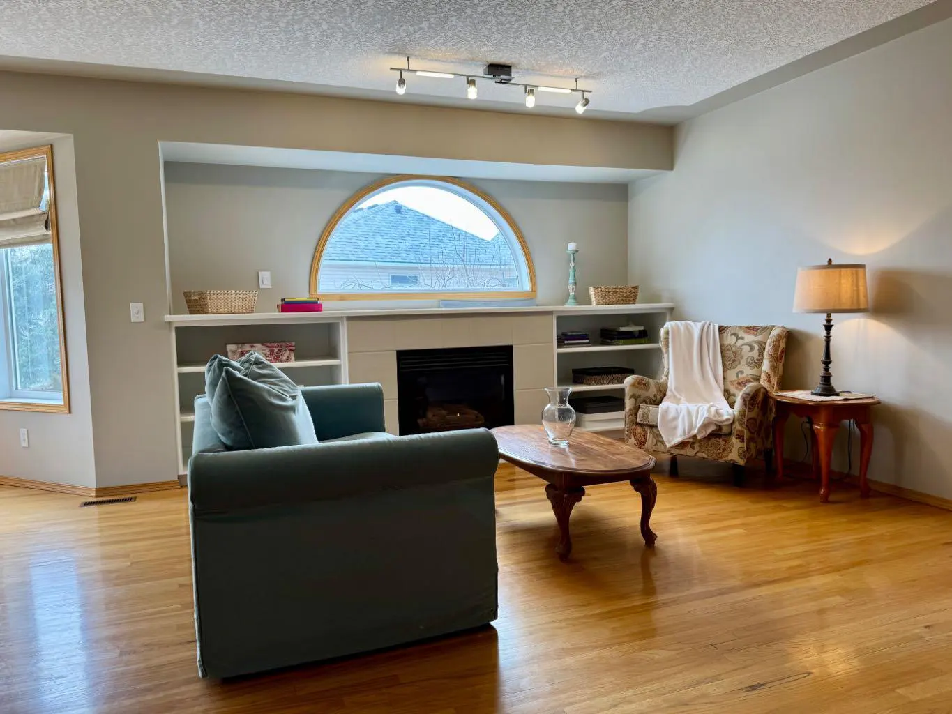 Living room with hardwood floors, a teal sofa, and a fireplace with built-in shelves. A patterned armchair and wood coffee table are also visible.