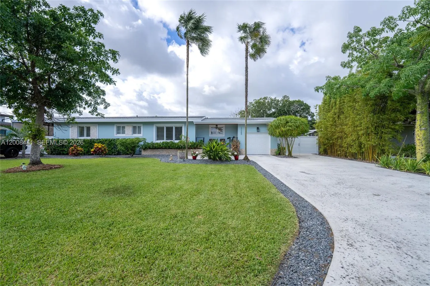 Light blue single-story house with a green lawn, palm trees, and a concrete driveway.