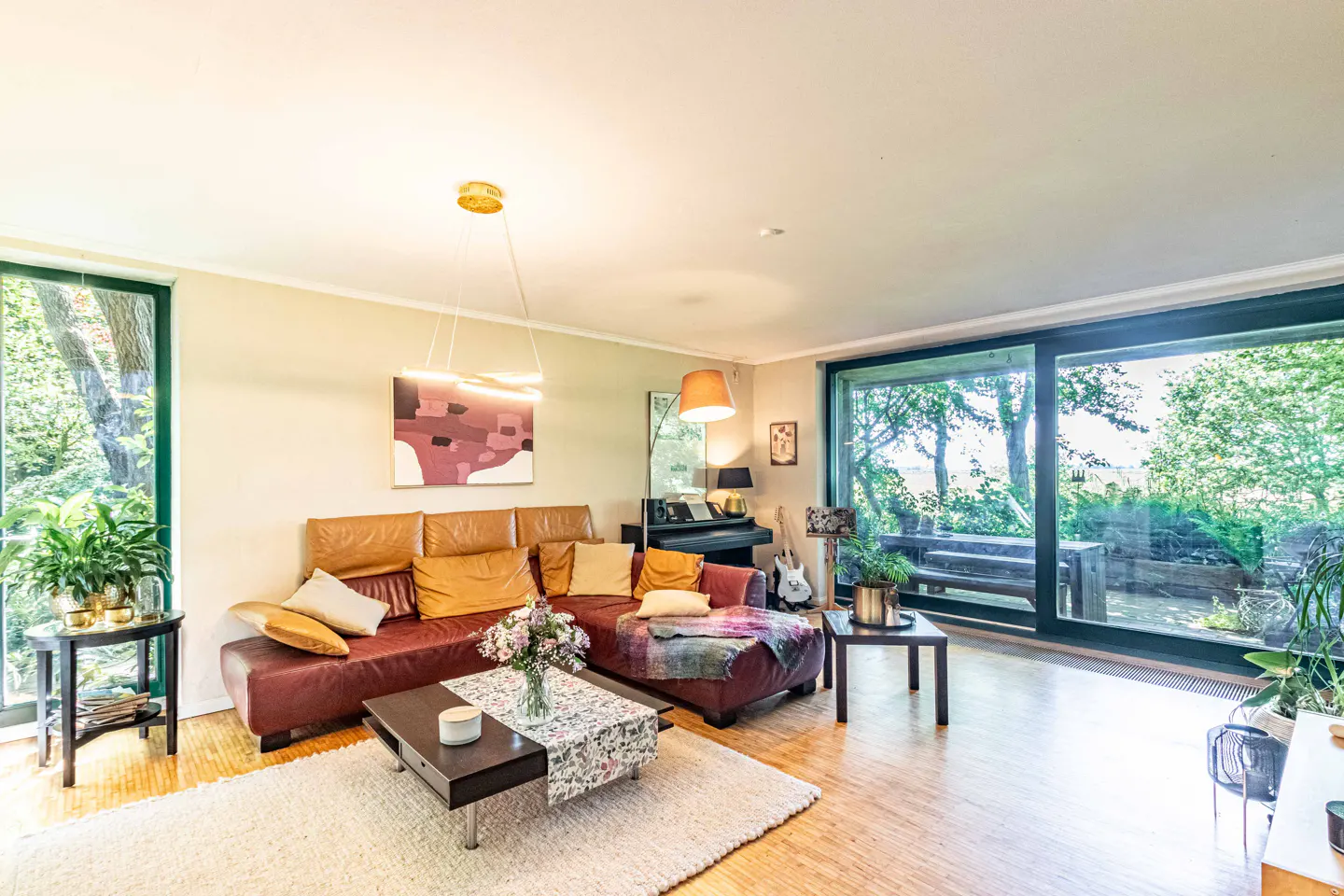 Bright living room with a dark red sectional sofa, coffee table, piano, and large windows overlooking a garden.
