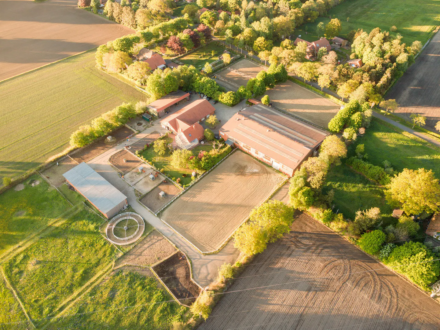 Aerial view of a horse farm with red-roofed buildings, sandy paddocks, green fields, and surrounding trees.