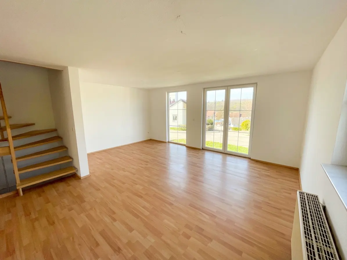 Bright, empty room with wood floors, white walls, and a staircase. Double doors lead to an outdoor view.