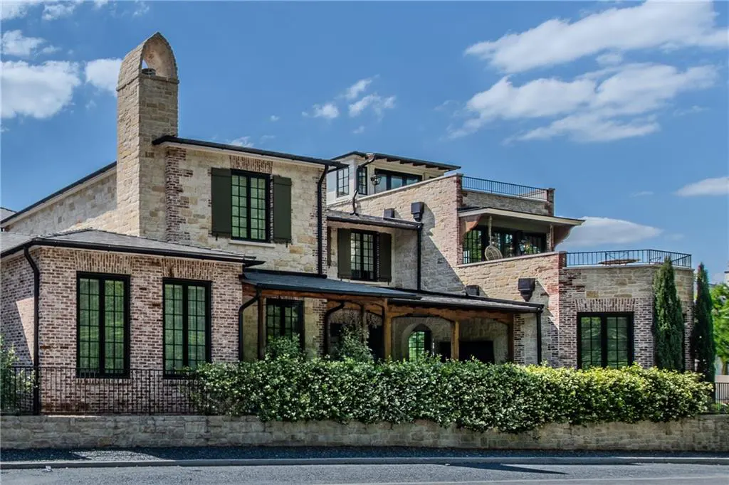 A multi-story stone and brick house with dark window frames, a tall chimney, and a manicured hedge under a blue sky.