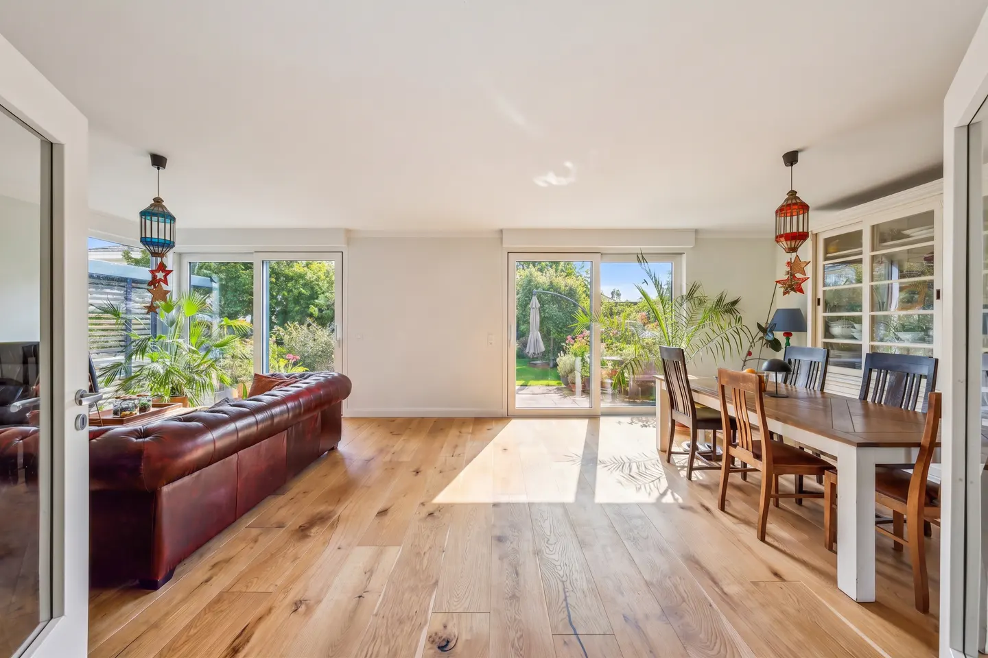 Bright living room with wood floors, a brown leather sofa, and a dining table with chairs. Sliding glass doors lead to a lush green garden.