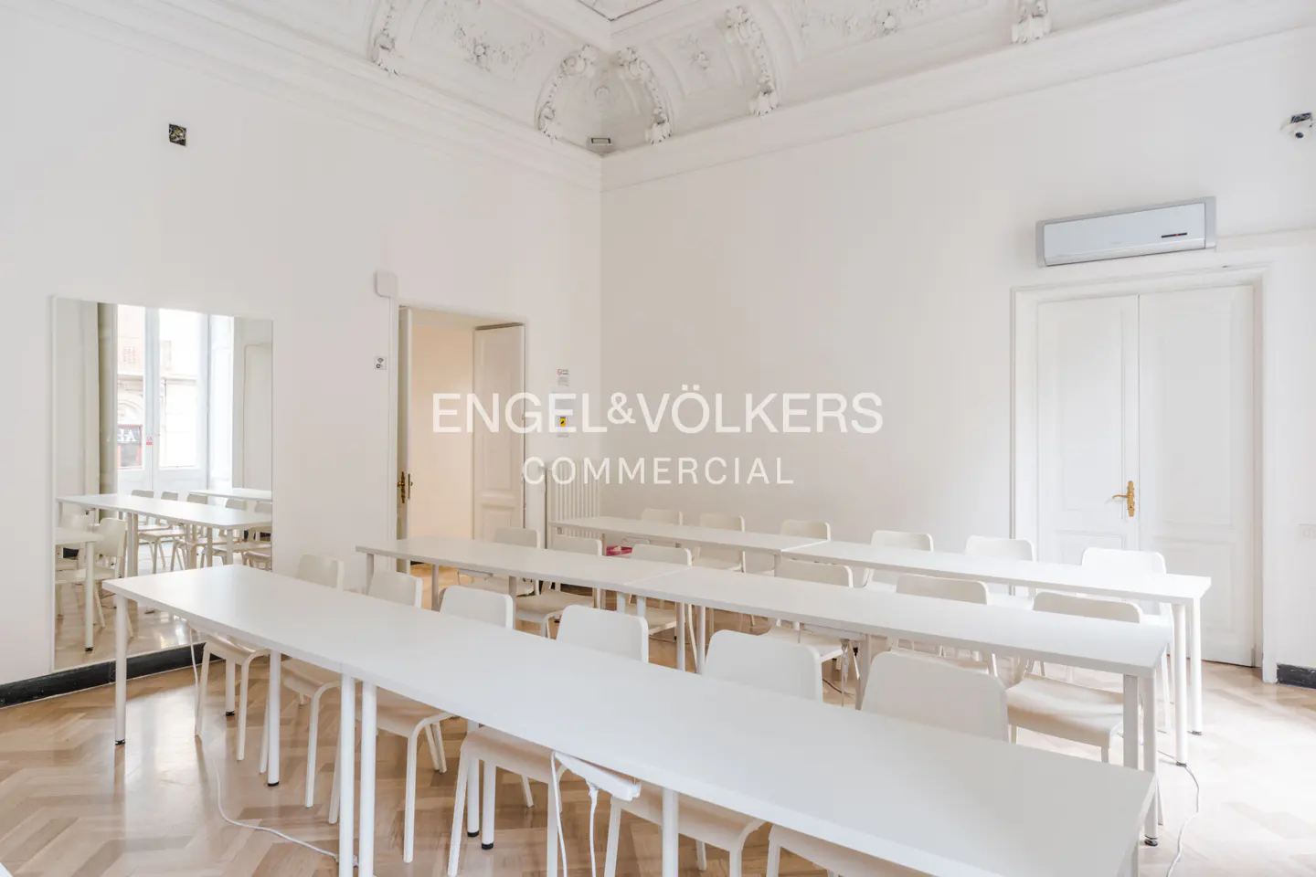 Bright, empty classroom with white walls, herringbone floors, and rows of white tables and chairs. Ornate ceiling detail. Engel & Völkers logo.