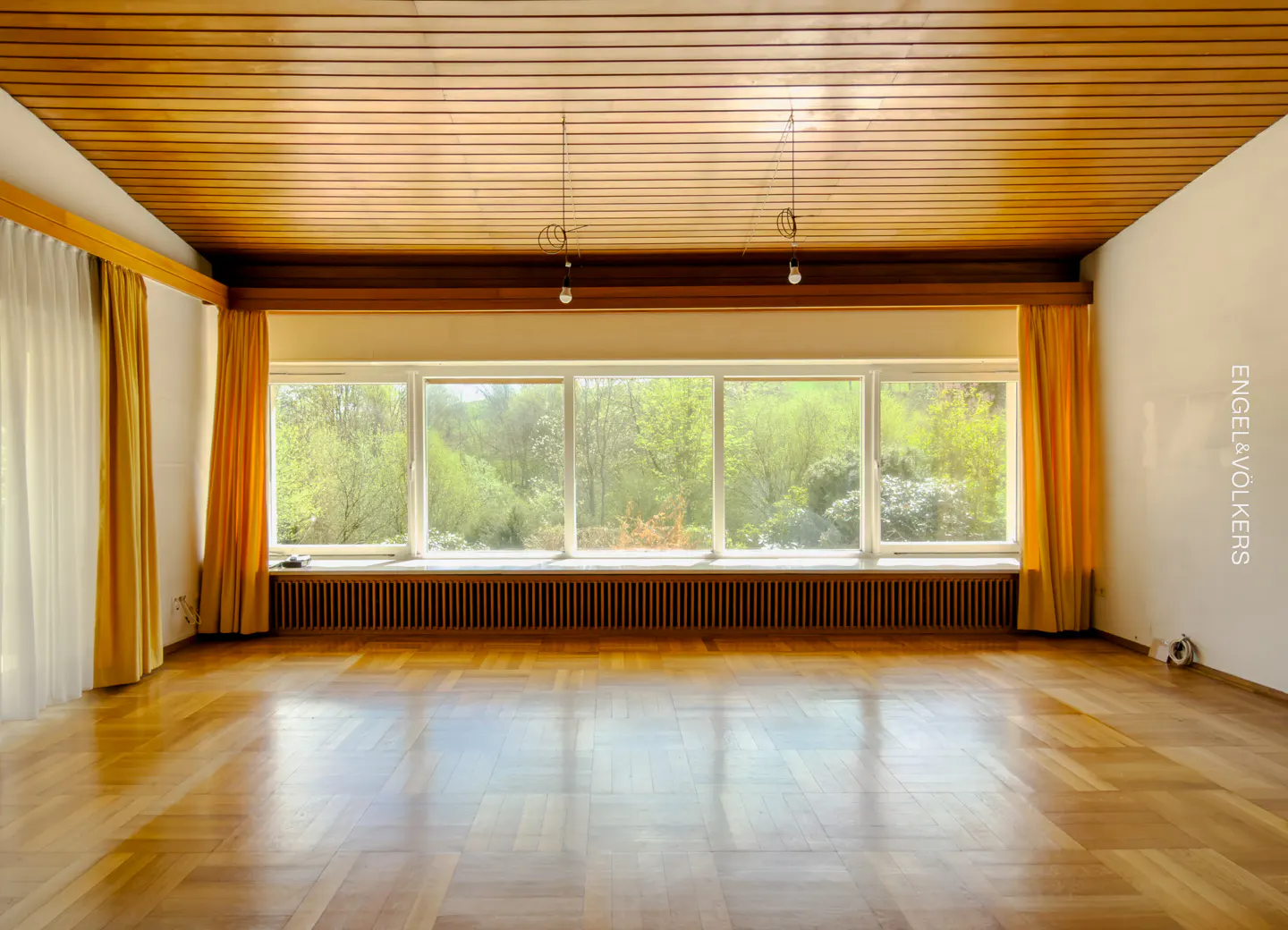 Empty room with wood floors, ceiling, and large window showing trees. Yellow curtains frame the window.