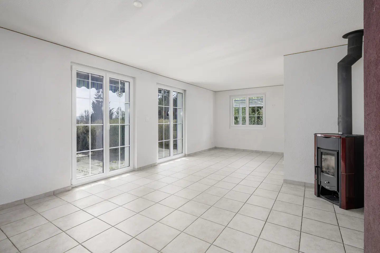 Bright, empty room with white walls, tile floor, and a wood-burning stove. Two sets of glass doors and a window offer outside views.