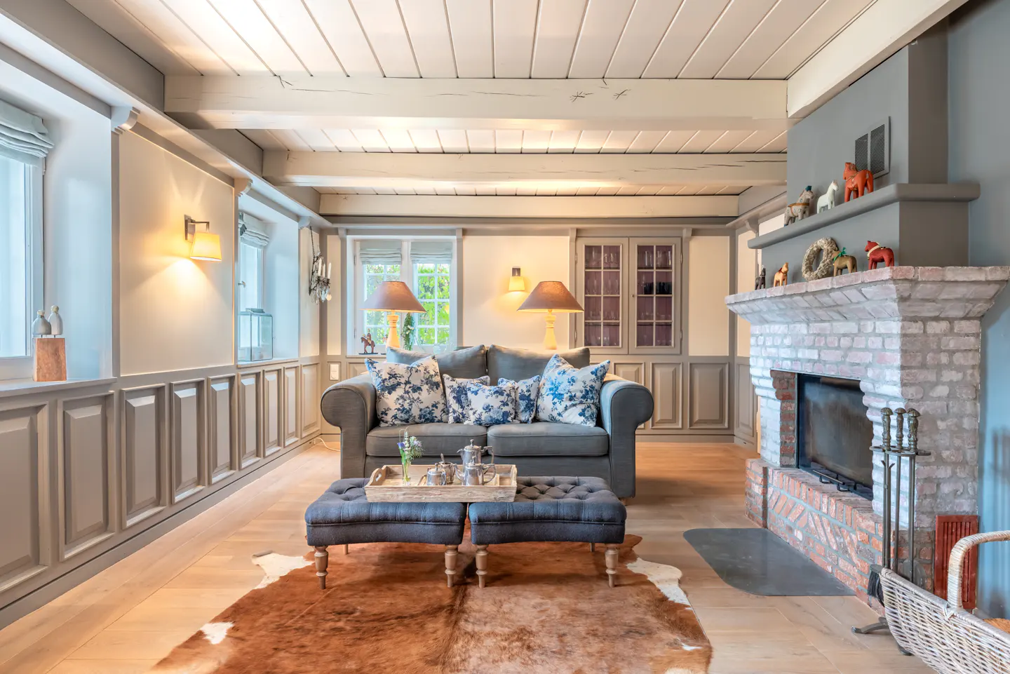 Living room with gray sofa, blue floral pillows, and ottoman on a brown cowhide rug. A brick fireplace is on the right.