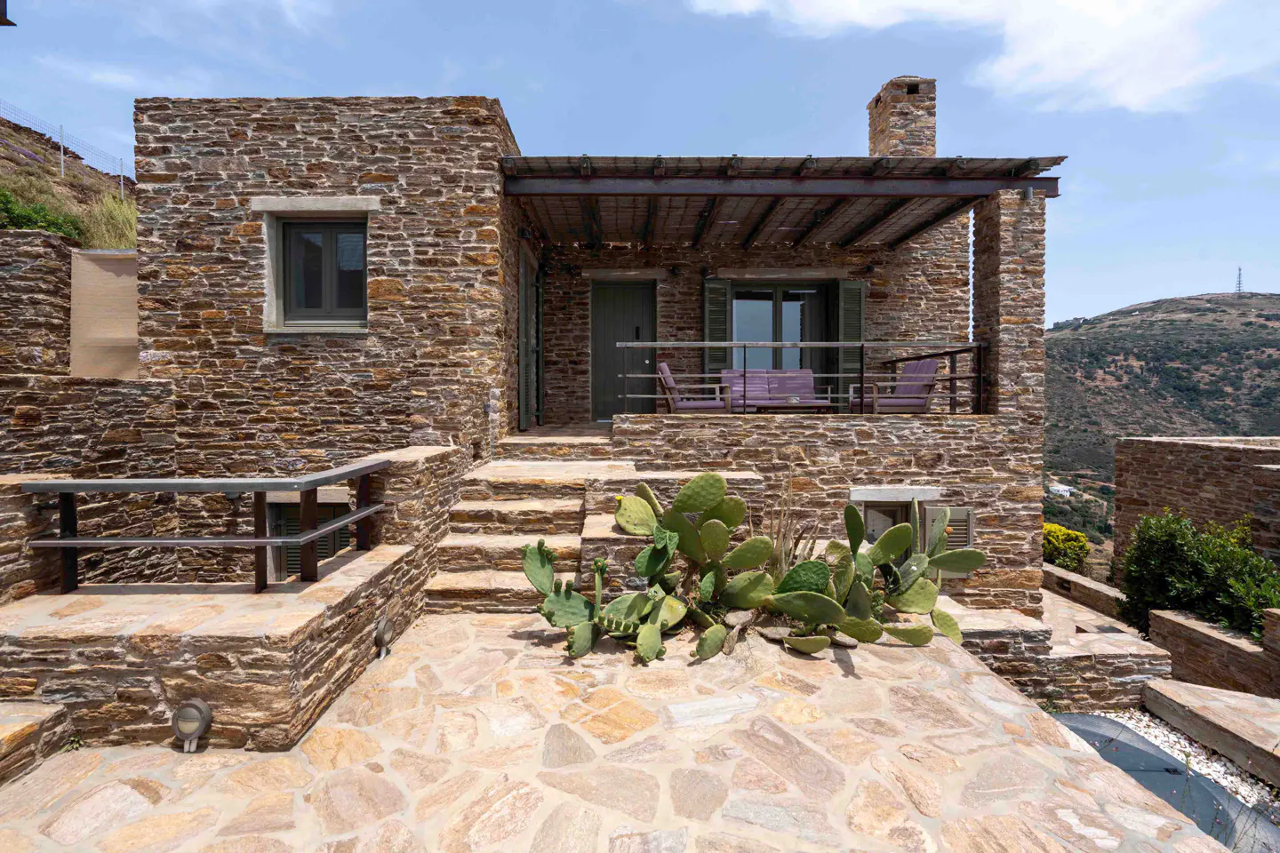 Rustic stone house with a patio, purple outdoor furniture, and a cactus garden against a mountainous landscape.
