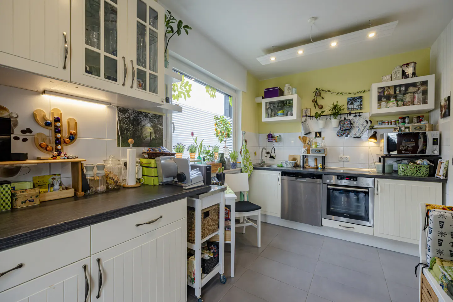 Bright kitchen with white cabinets, gray countertops, and gray tile floor. Window with plants above sink. Appliances include oven, dishwasher, and microwave.