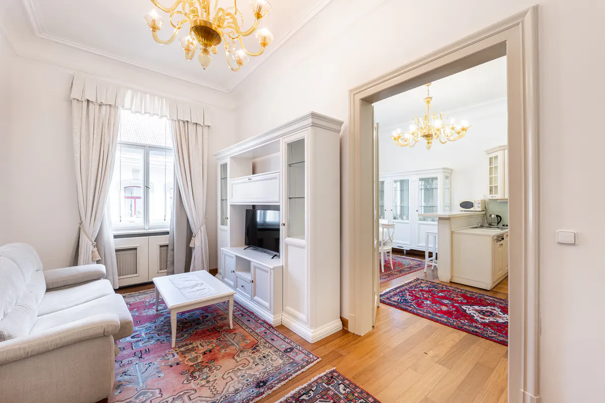 Living room with white sofa, TV cabinet, and coffee table on a patterned rug. A doorway leads to a kitchen with white cabinets and a dining area. A chandelier hangs in both rooms.