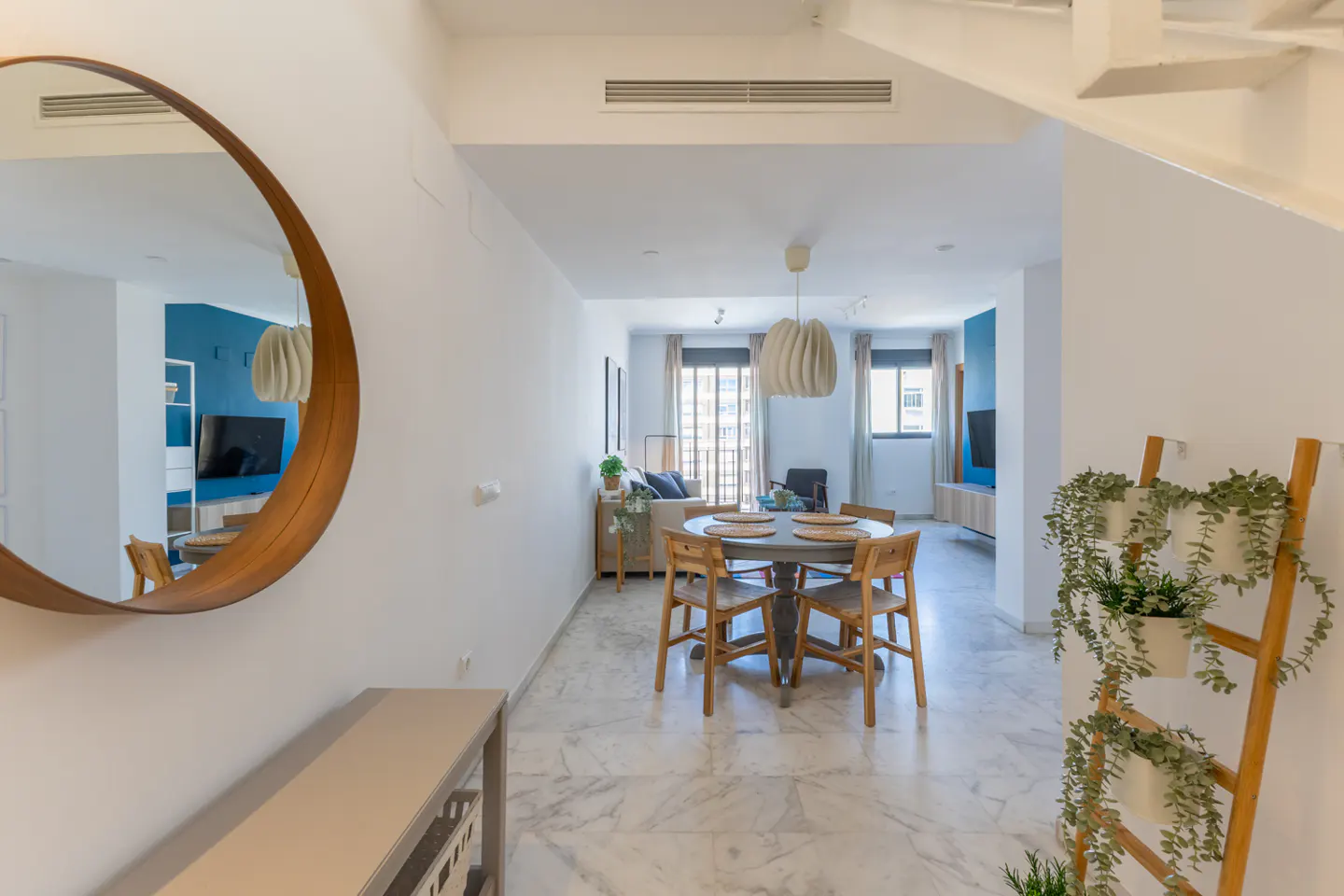 Bright, modern apartment interior with a round wooden mirror, dining table, and plant ladder. White walls and marble floors create a clean, airy feel.