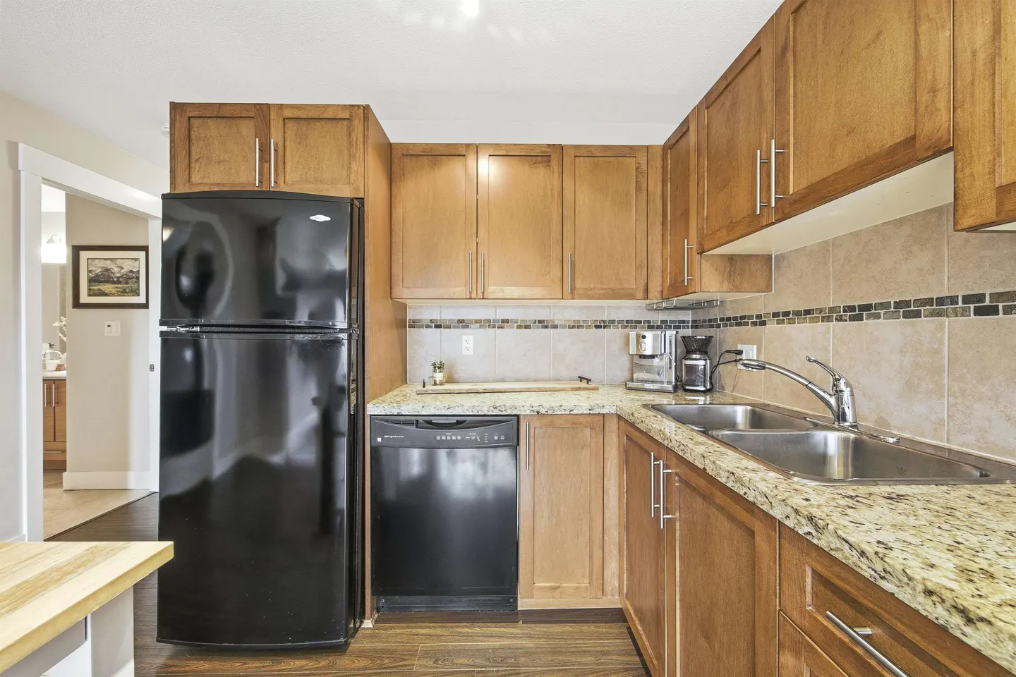A kitchen with wood cabinets, granite countertops, and a black refrigerator and dishwasher. A stainless steel sink and coffee maker are also visible.