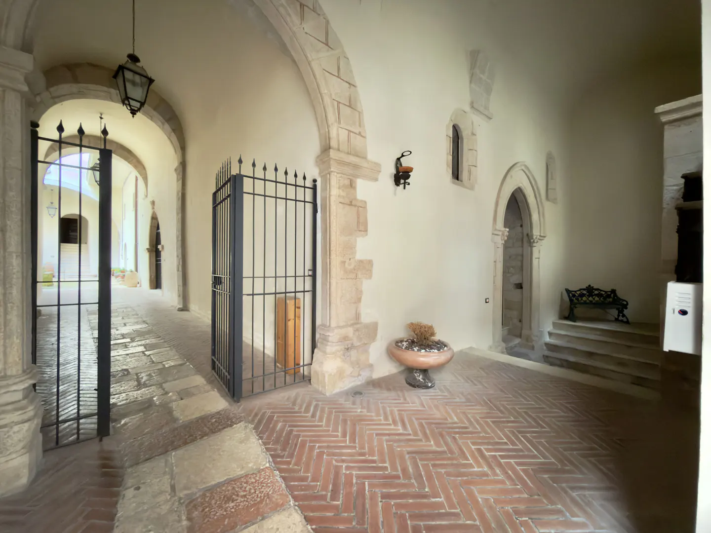 Courtyard with brick floors, stone arches, and black iron gates. A bench sits near a stone doorway. A potted plant adds a touch of greenery.