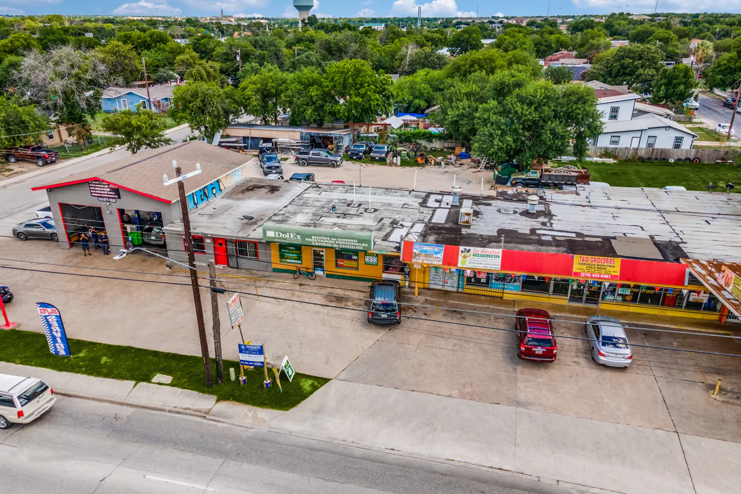 Aerial view of a commercial property with a gray auto shop, yellow DolEx, and a red-roofed grocery store. Cars are parked in front, with green trees and a water tower in the background.