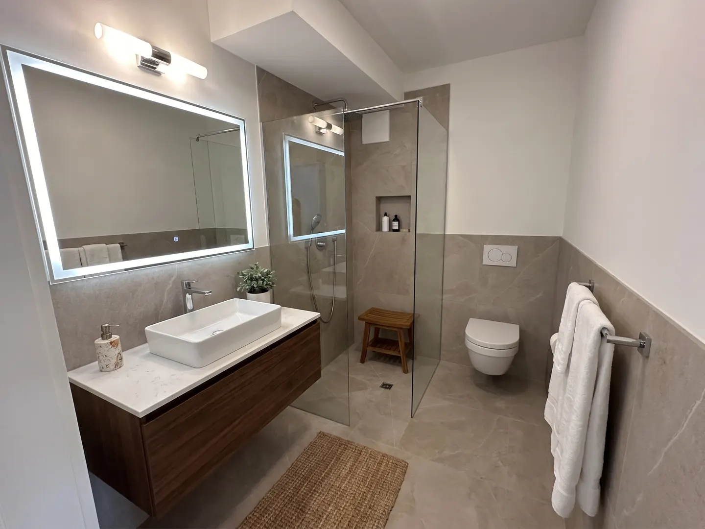 Modern bathroom with a wood vanity, white sink, and illuminated mirror. A glass shower, toilet, and towel rack are visible.