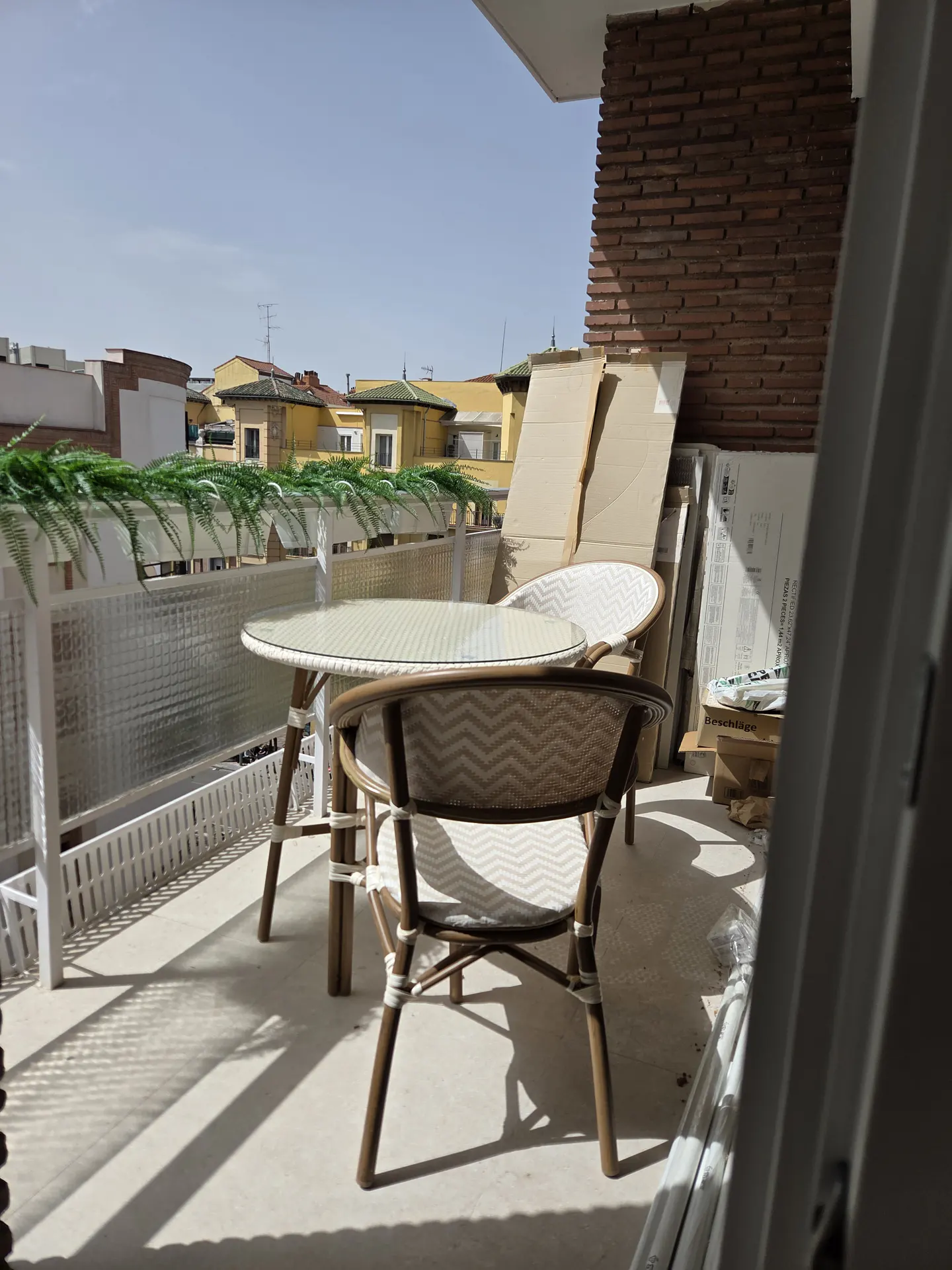 A balcony with a round glass table and two woven chairs, overlooking a sunny urban neighborhood with yellow buildings.