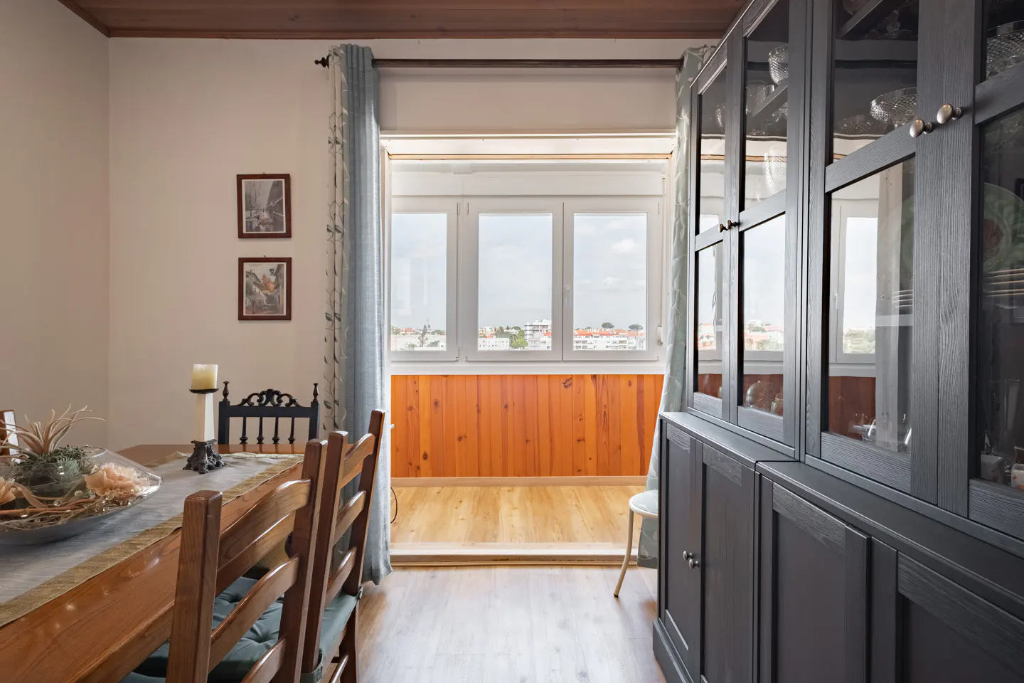 Dining room with wooden table, dark grey hutch, and a view through a window to a balcony with wood-paneled walls.