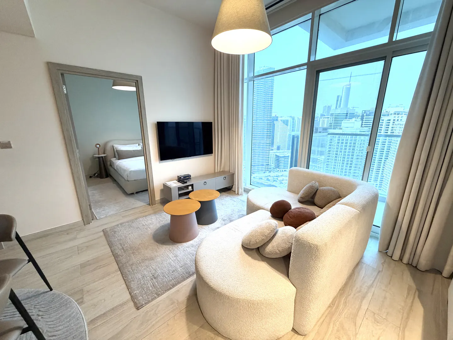 Living room with a curved white sofa, city view through large windows, and a glimpse of a bedroom through a doorway.