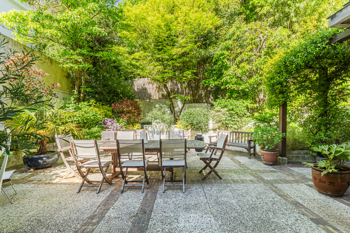 Outdoor patio with stone flooring, surrounded by lush green trees and plants. A wooden table and chairs sit in the center.