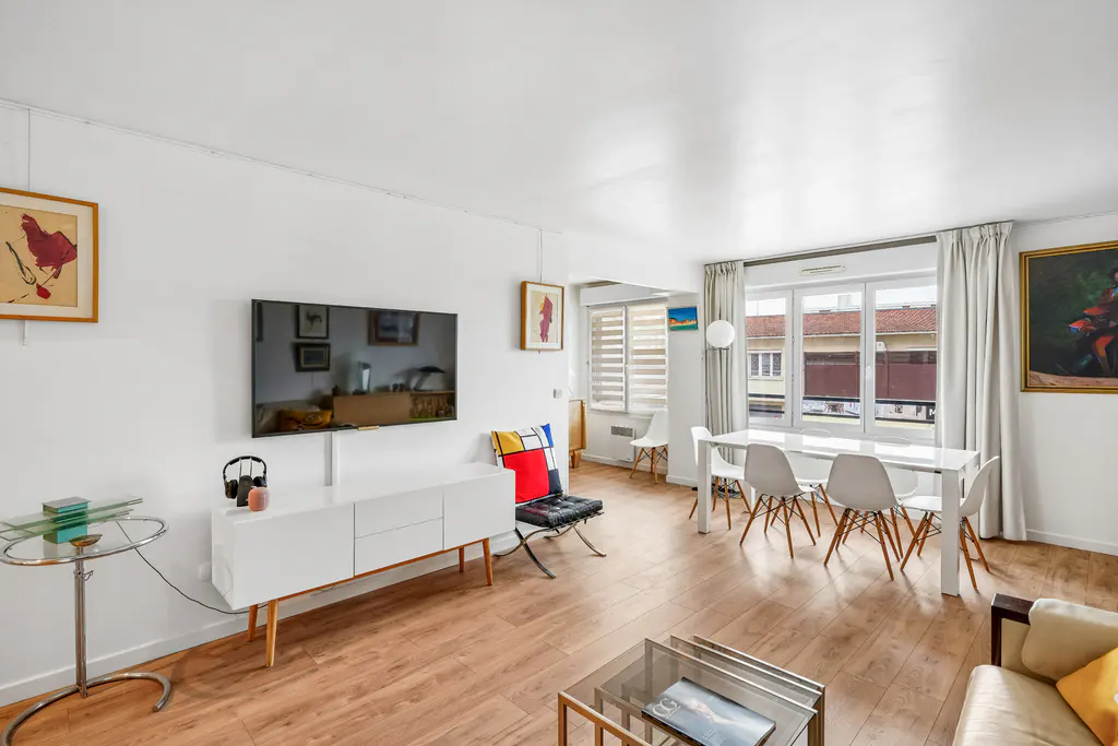 Bright living room with white walls, wood floors, and modern furniture. A TV hangs above a white console table. Dining area with white table and chairs near a window.