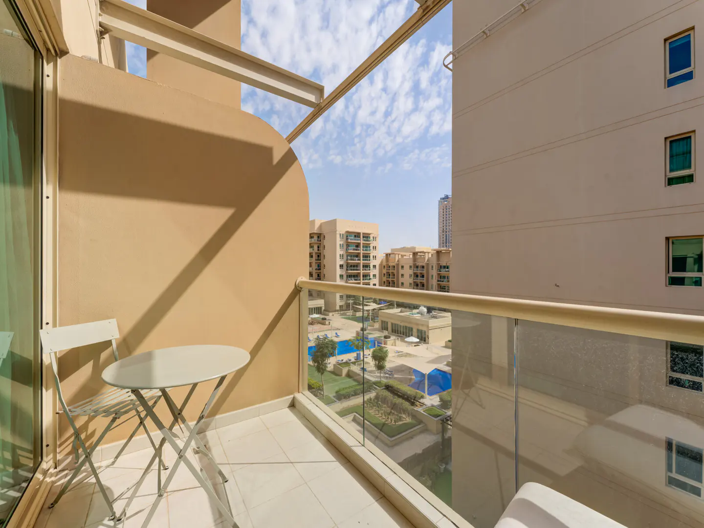 Balcony view with a small white table and chair, overlooking a courtyard with pools and buildings under a blue sky.