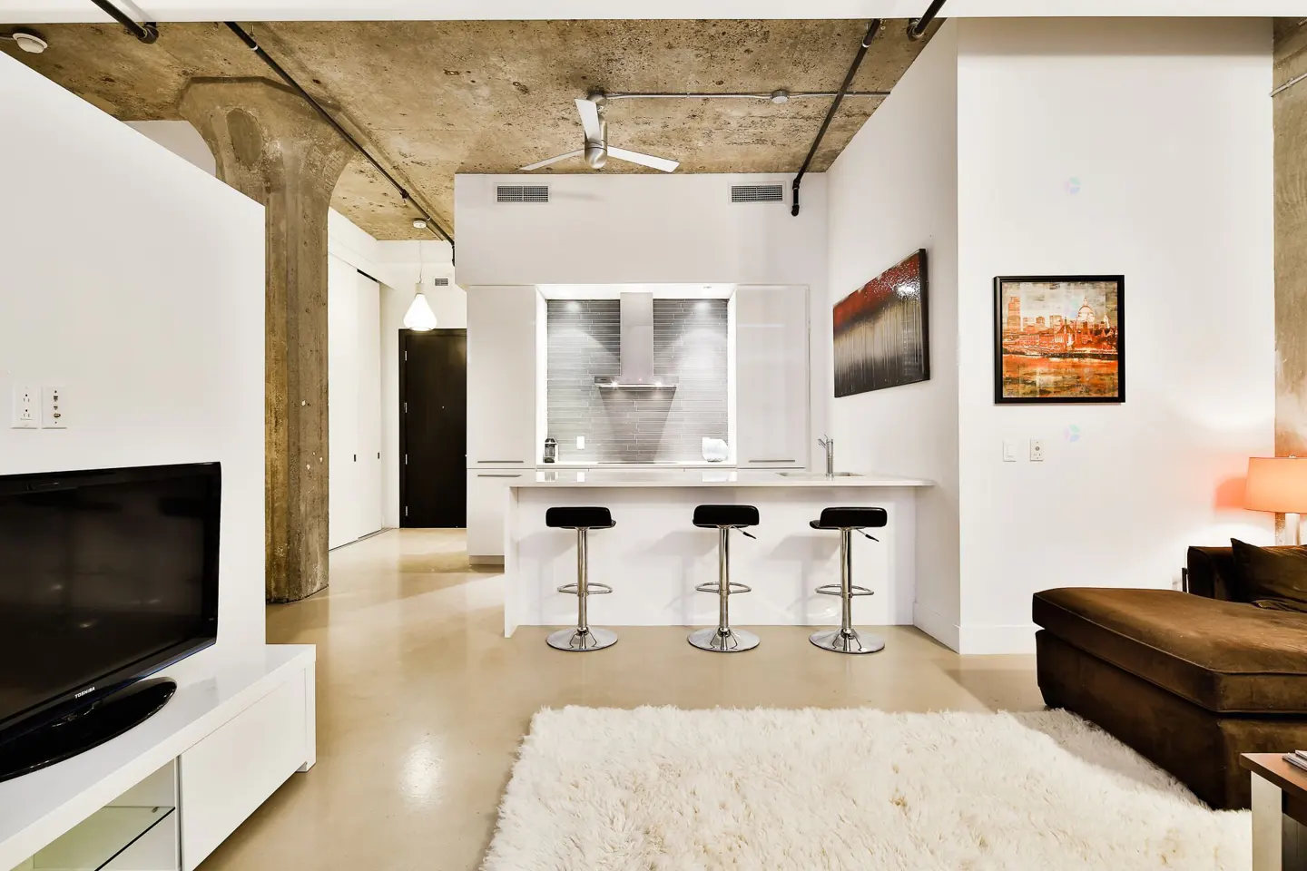 Modern loft living room with concrete ceiling, white kitchen island with three stools, and a brown sofa on a white rug.