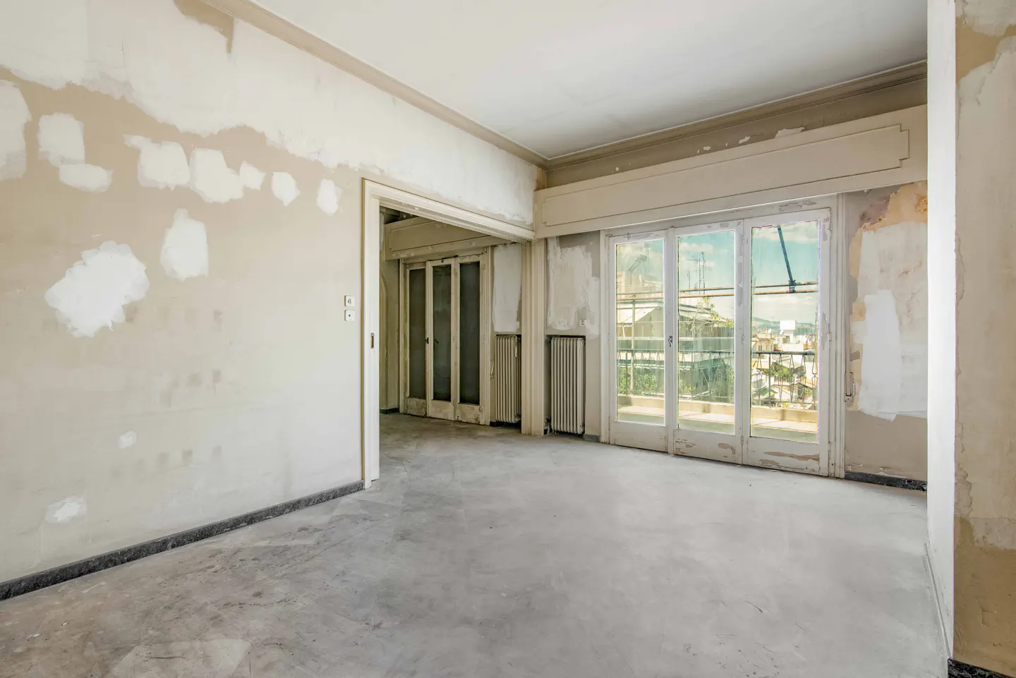 Unfinished room with patched walls, concrete floor, and white-framed glass doors leading to a balcony with a city view.