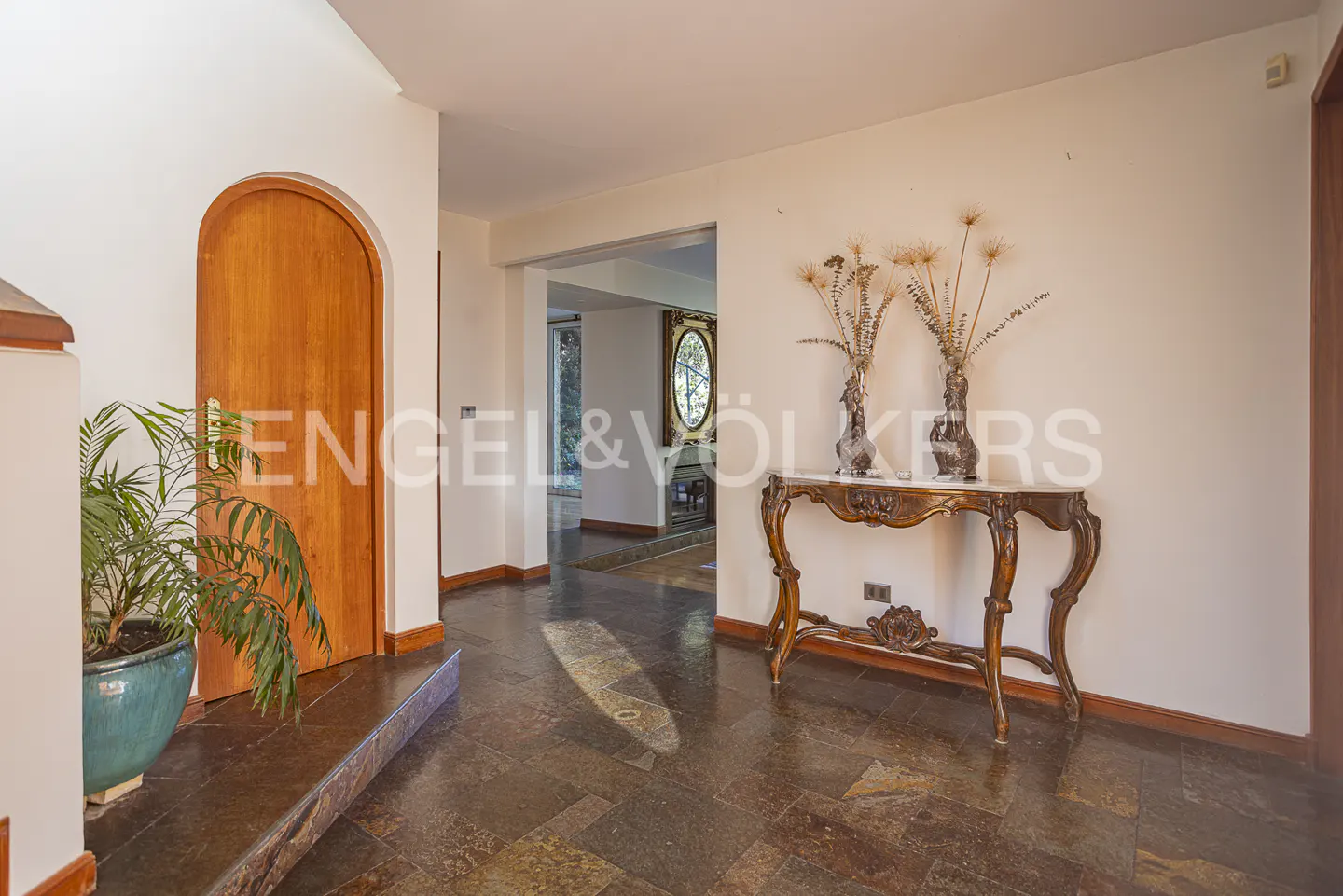Bright foyer with dark stone tile floors, an arched wooden door, a potted plant, and an ornate console table with vases.