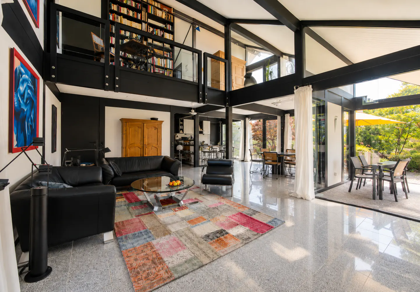 Modern living room with black leather sofas, a colorful patchwork rug, and a mezzanine library overlooking the space.