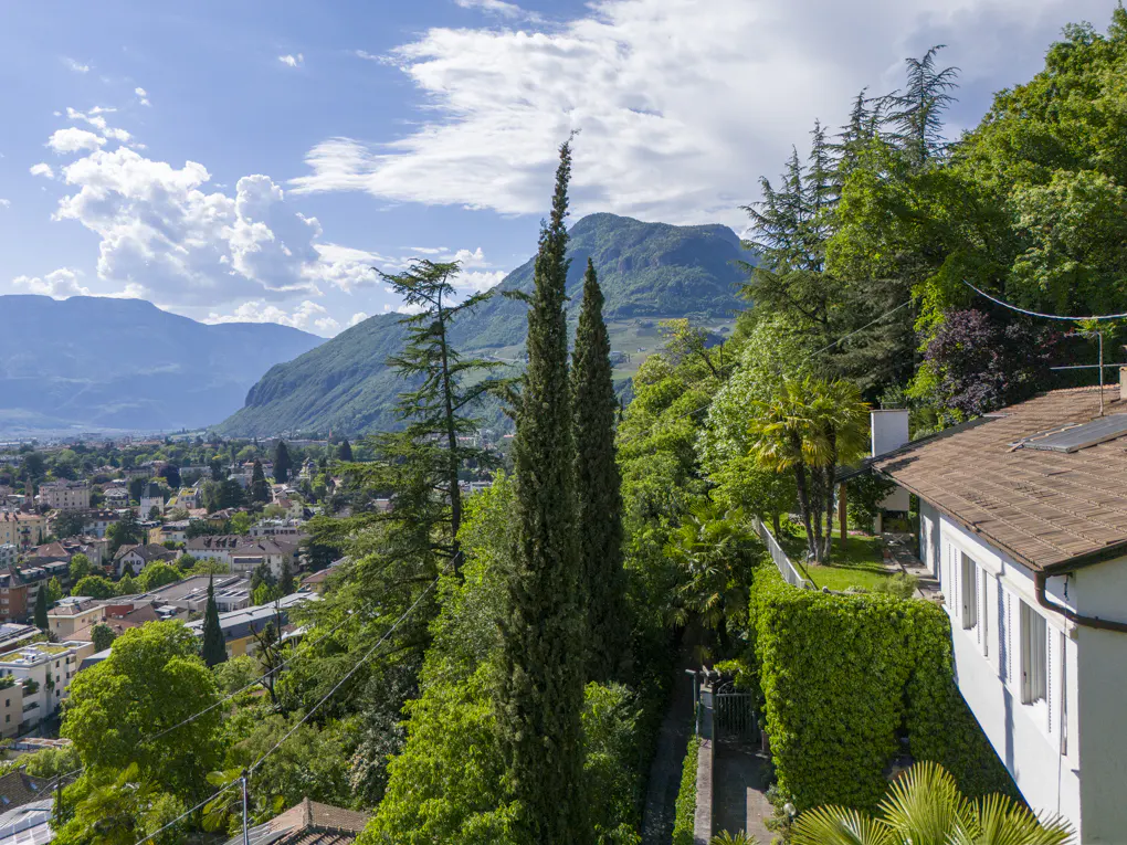 Scenic view of a town nestled in a valley, with mountains, lush trees, and a white house with a brown roof in the foreground.