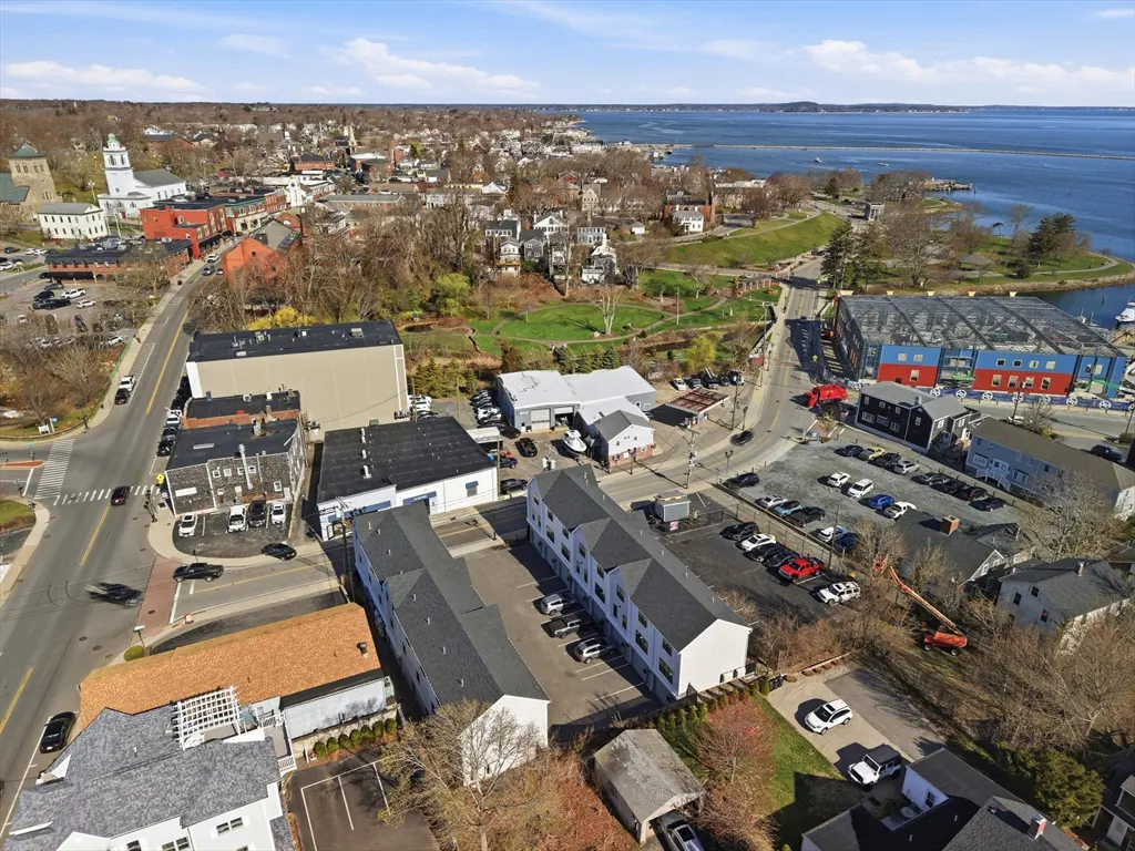 Aerial view of a coastal town with buildings, roads, parked cars, and a harbor with blue water under a partly cloudy sky.