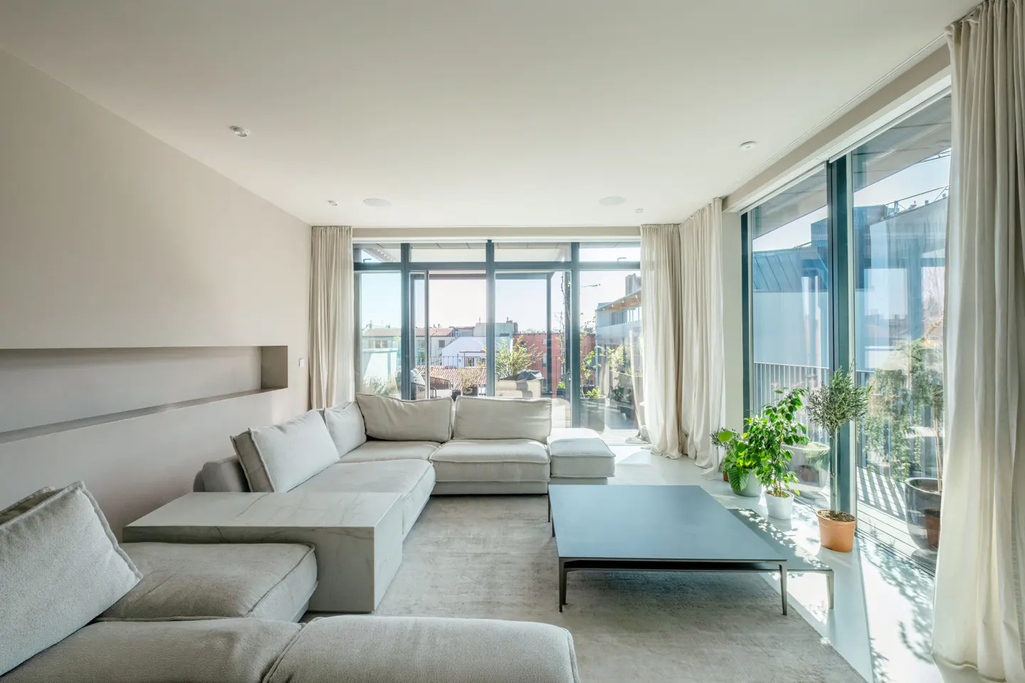 Modern living room with a large beige sectional sofa, low coffee table, and floor-to-ceiling windows overlooking a city.
