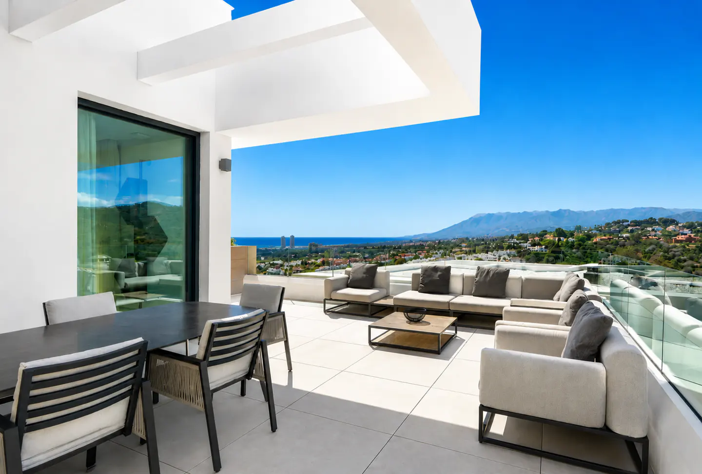 Modern white patio with dining table, chairs, and outdoor sofa overlooking a scenic view of mountains and the sea under a clear blue sky.