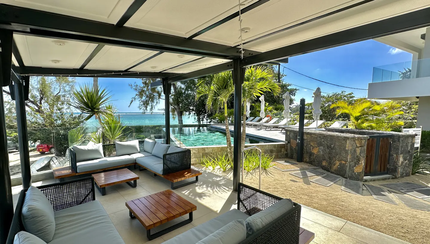 Outdoor lounge area with gray sofas, wooden tables, and a black pergola. A pool and ocean are visible in the background.