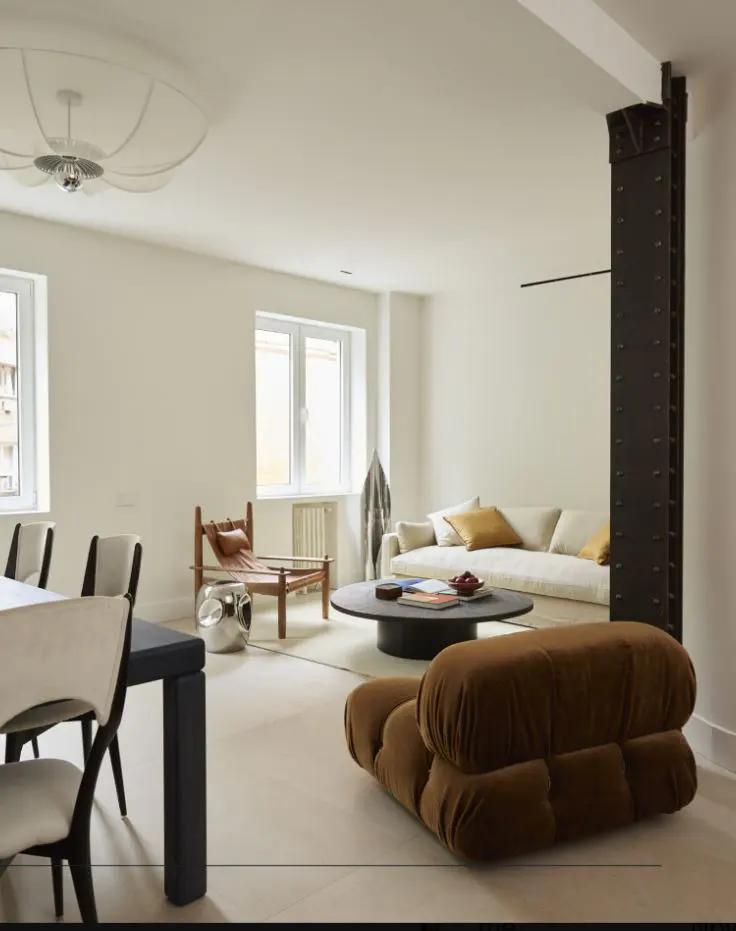 Bright, modern living room with white walls, large windows, and a mix of seating including a brown floor cushion and a cream sofa.