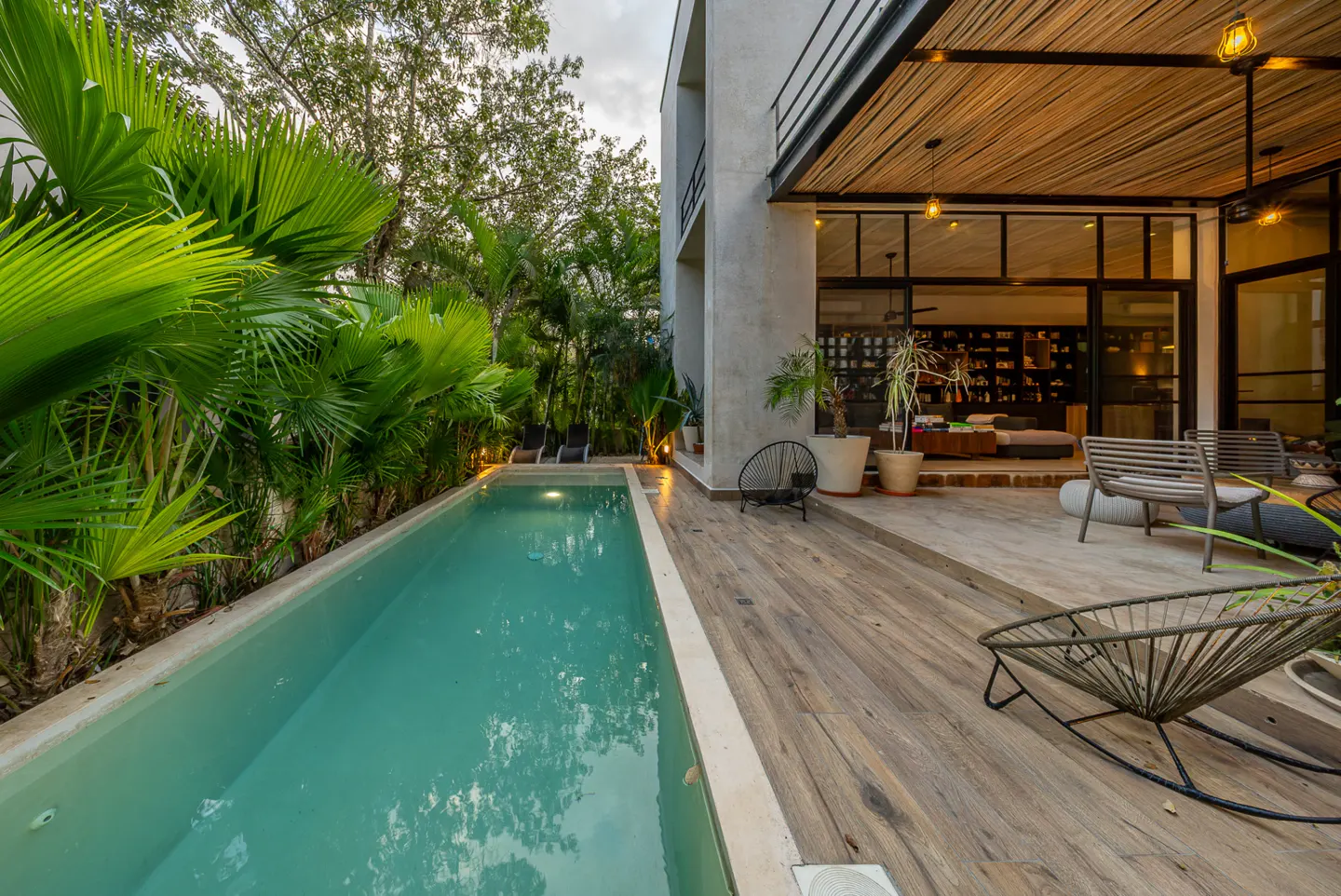 Outdoor view of a modern house with a pool, wooden deck, and tropical plants. The house has large glass doors and a bamboo-covered porch.