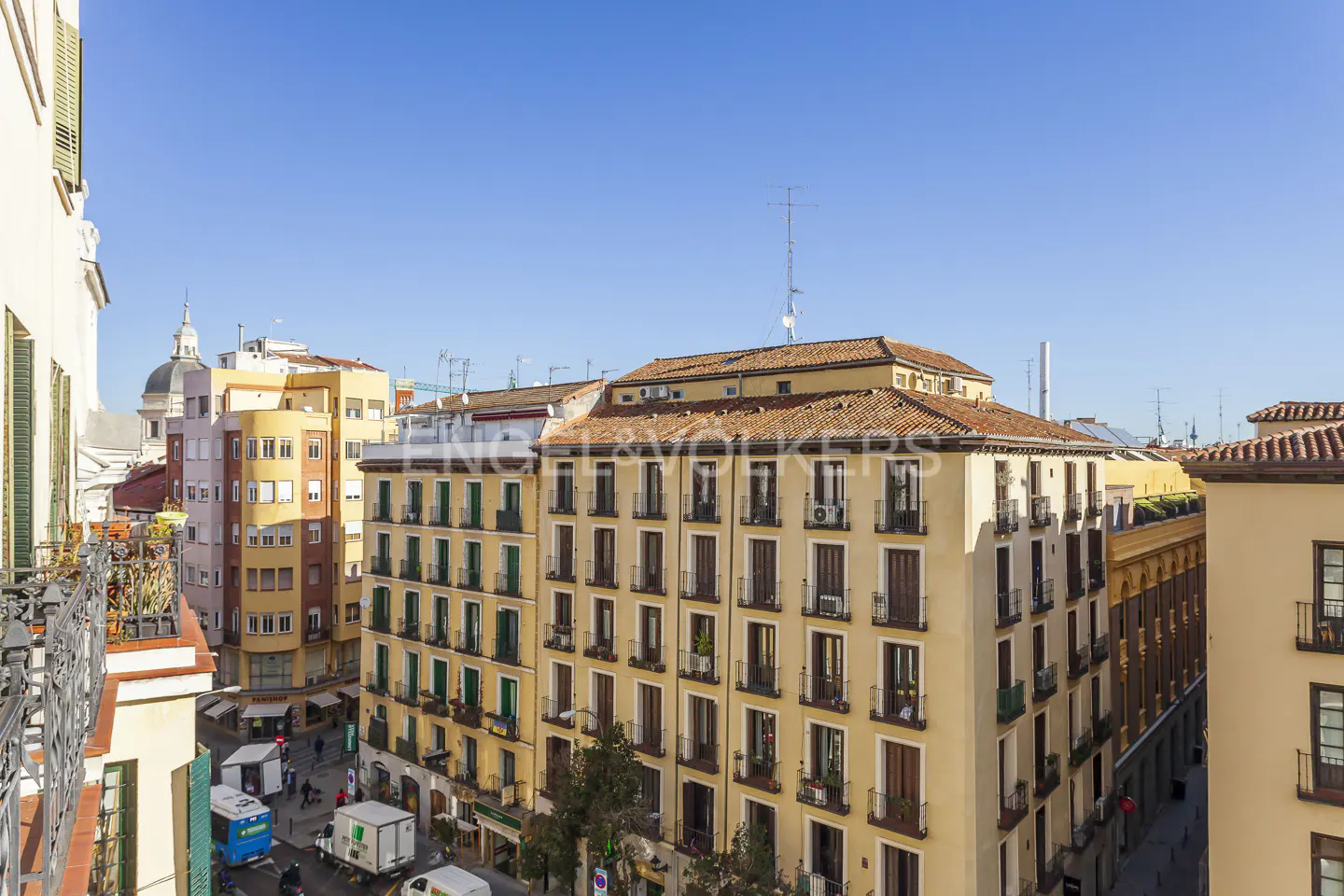Cityscape view of yellow buildings with brown trim under a clear blue sky. Balcony with plants in the foreground.