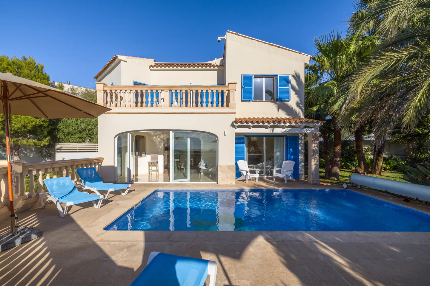Exterior of a two-story beige house with a blue pool, lounge chairs, and palm trees.