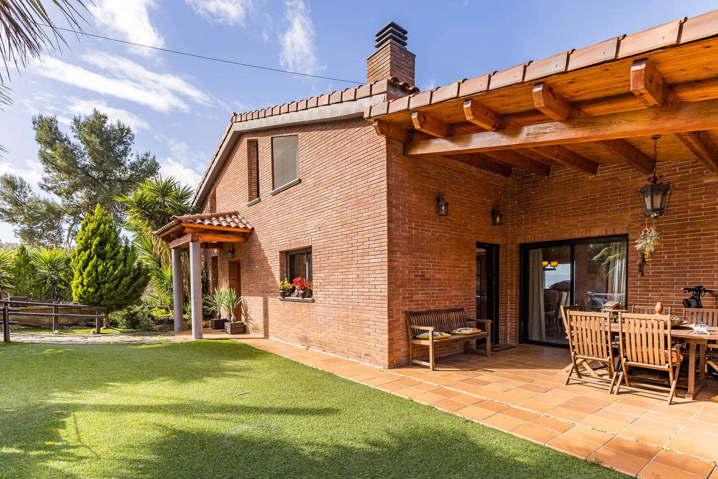 Red brick house with a tiled patio, wooden pergola, outdoor dining set, and a green lawn under a bright blue sky.