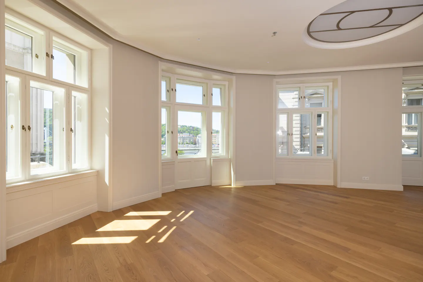 Bright, empty room with hardwood floors, white walls, and large windows offering city views. Sunlight streams in, creating shadows on the floor.