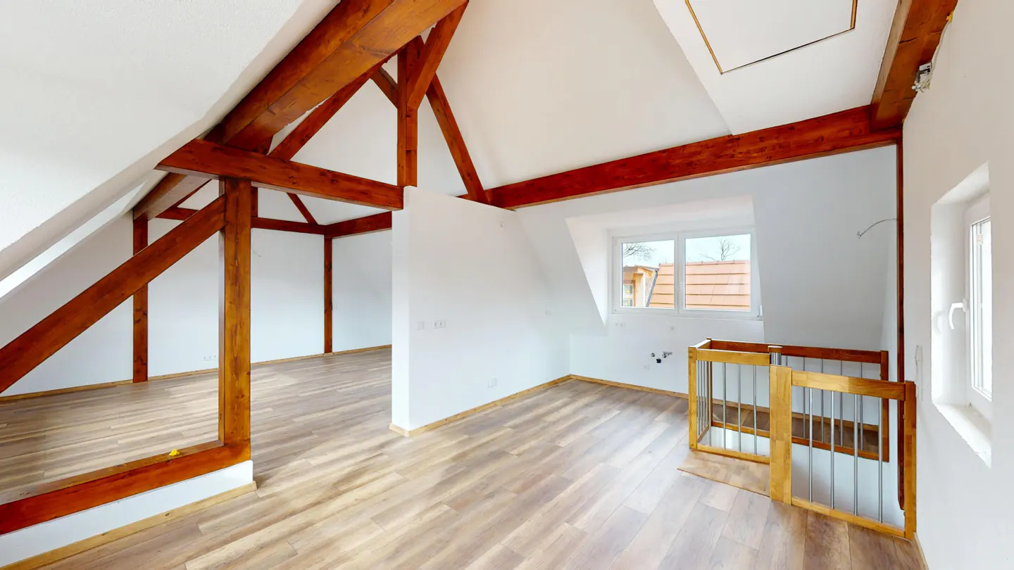 Attic room with wood floors, white walls, and exposed brown wood beams. A window and a wooden safety gate are visible.