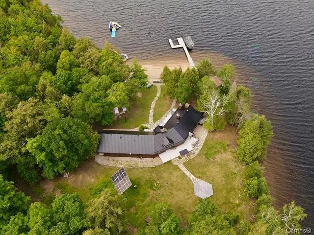 Aerial view of a lake house with a black roof, surrounded by green trees and a dock on the water. Solar panels are visible on the lawn.