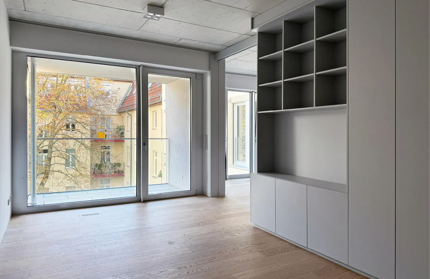 A bright, empty room with light wood floors, a gray built-in shelf and cabinet, and a large window with a view of a building and tree.