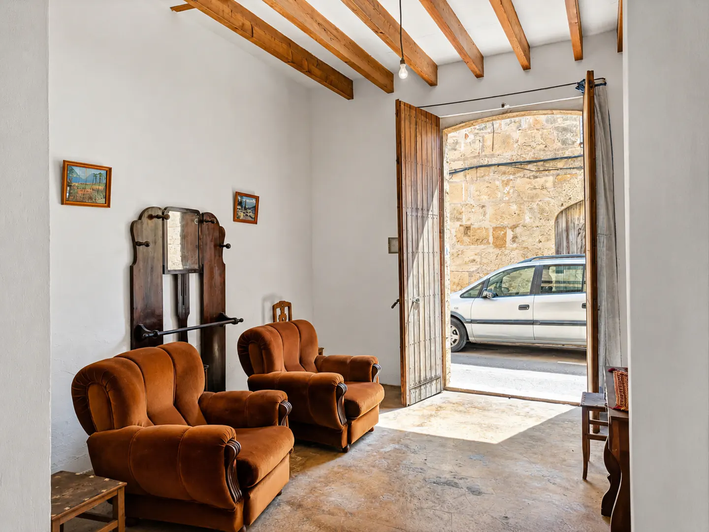 Living room with two brown armchairs, wooden beams on the ceiling, and an open door leading to a street with a parked car.