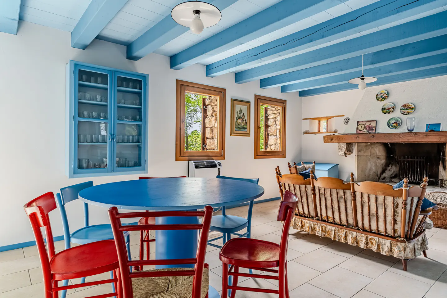 Bright dining room with blue table, red chairs, and blue ceiling beams. A fireplace and wooden sofa are in the background.