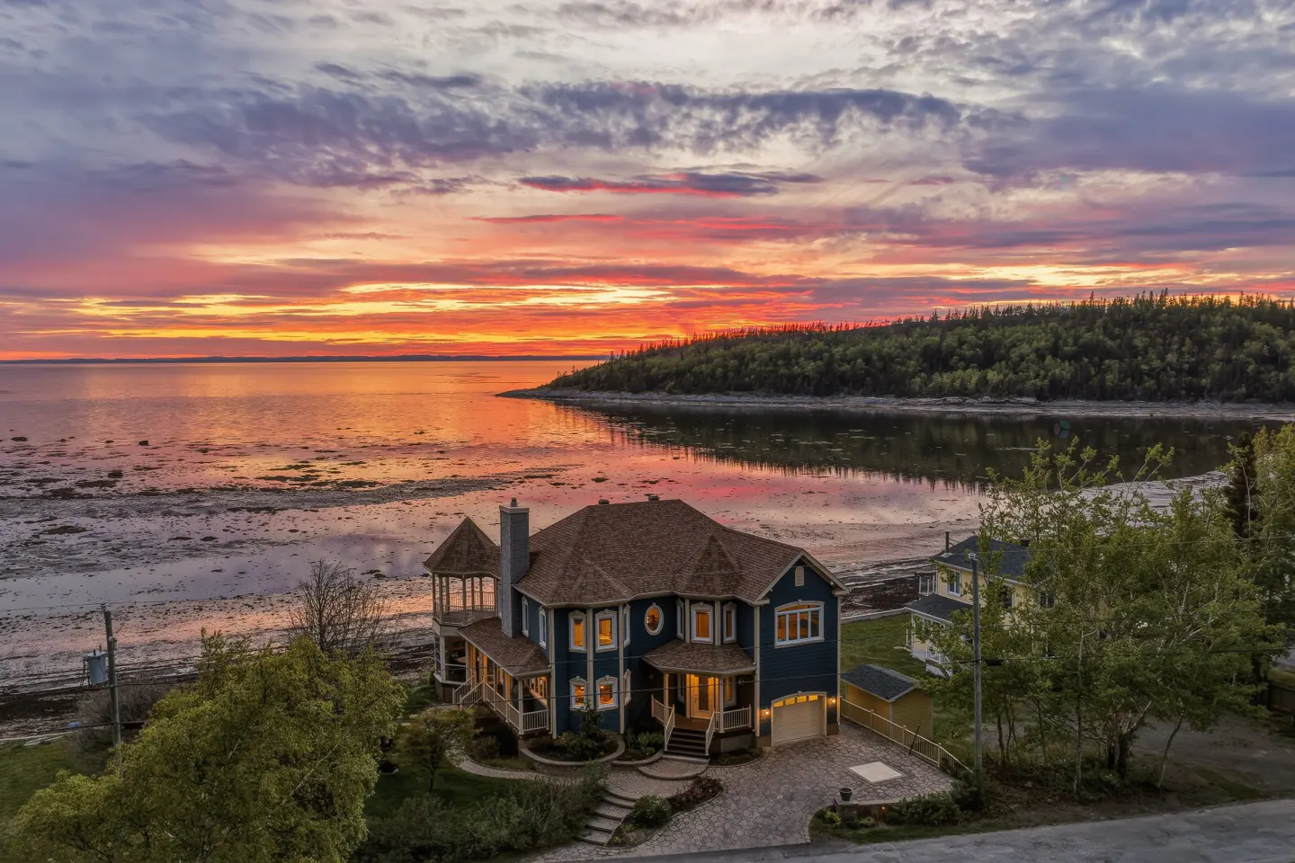 Aerial view of a blue house with a turret at sunset, overlooking a calm bay with a vibrant orange and purple sky.